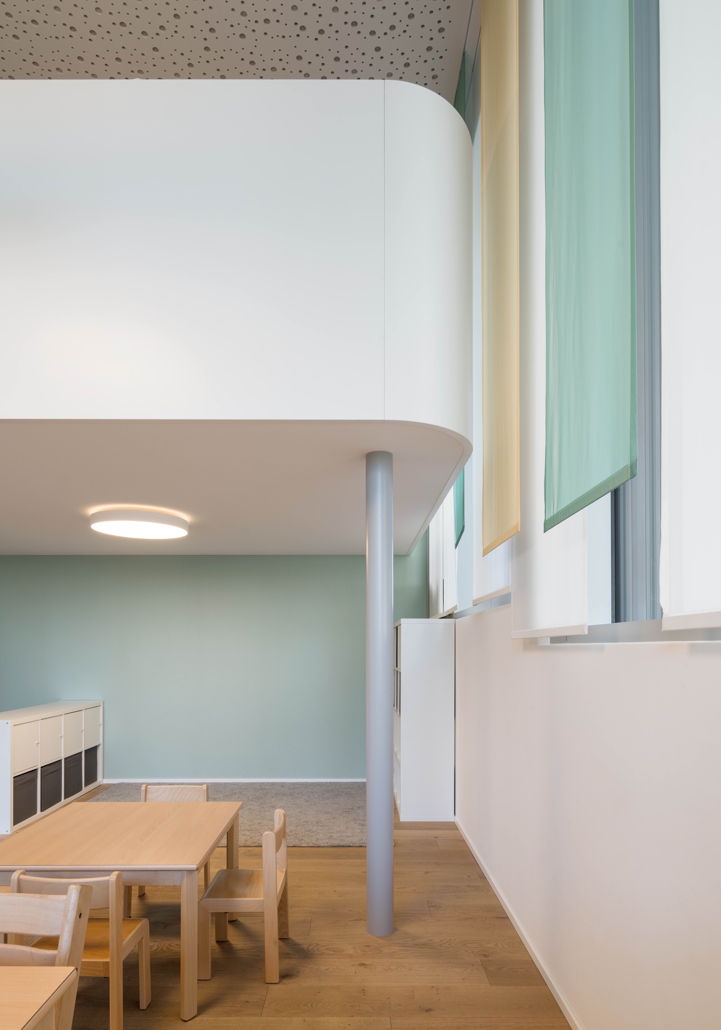 Innenraum eines modernen, hellen Raumes mit weißen Wänden, Holzfußboden, einem kleinen Tisch und Stühlen, großen Fenstern mit transparenten Vorhängen, einem runden Deckenleuchte und einer weißen Balkon- oder Mezzanine.