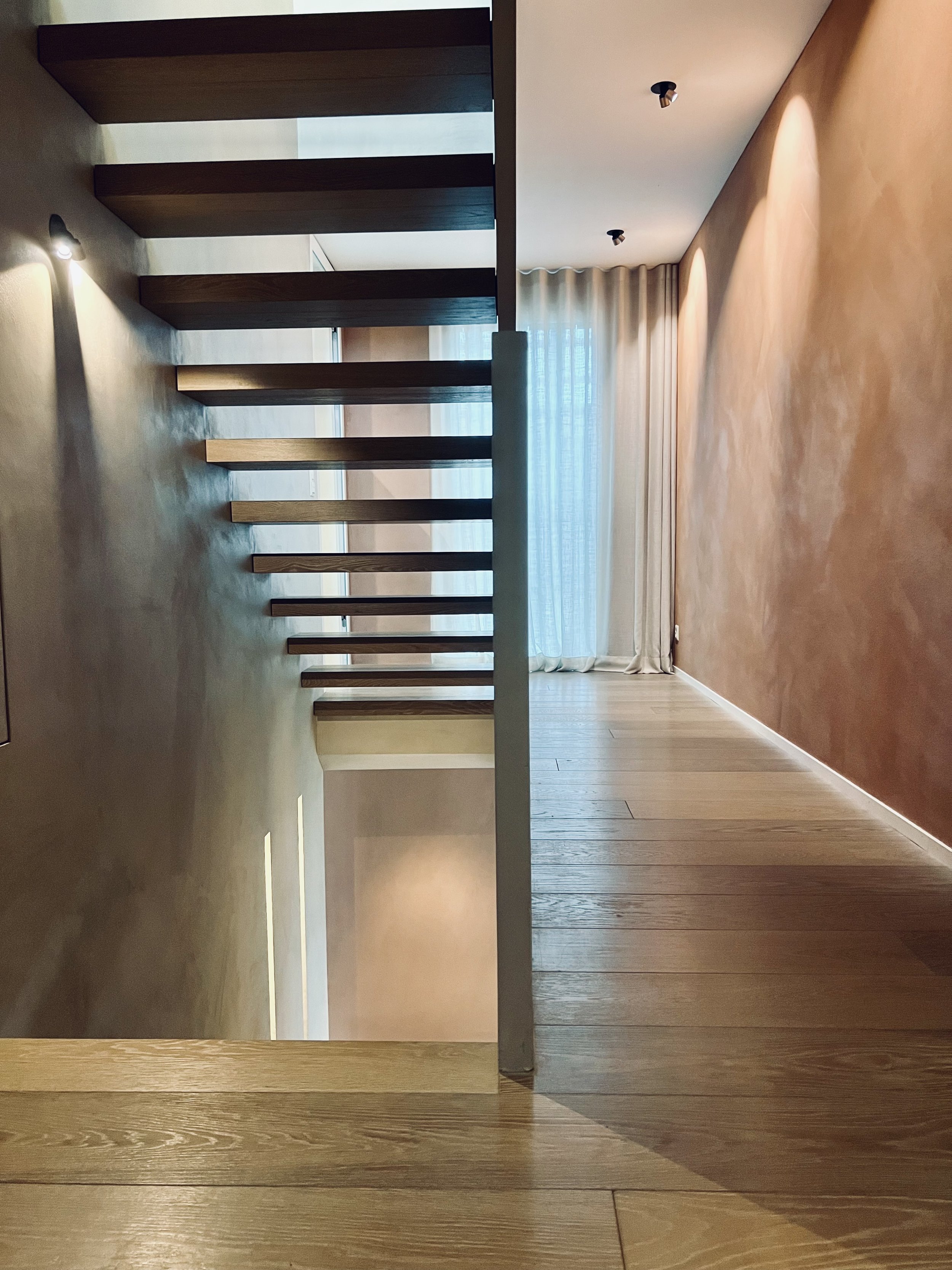 Innenaufnahme eines modernen Wohnhauses mit Holzboden, Treppe ohne Geländer und großen Fenstern mit Gardinen.