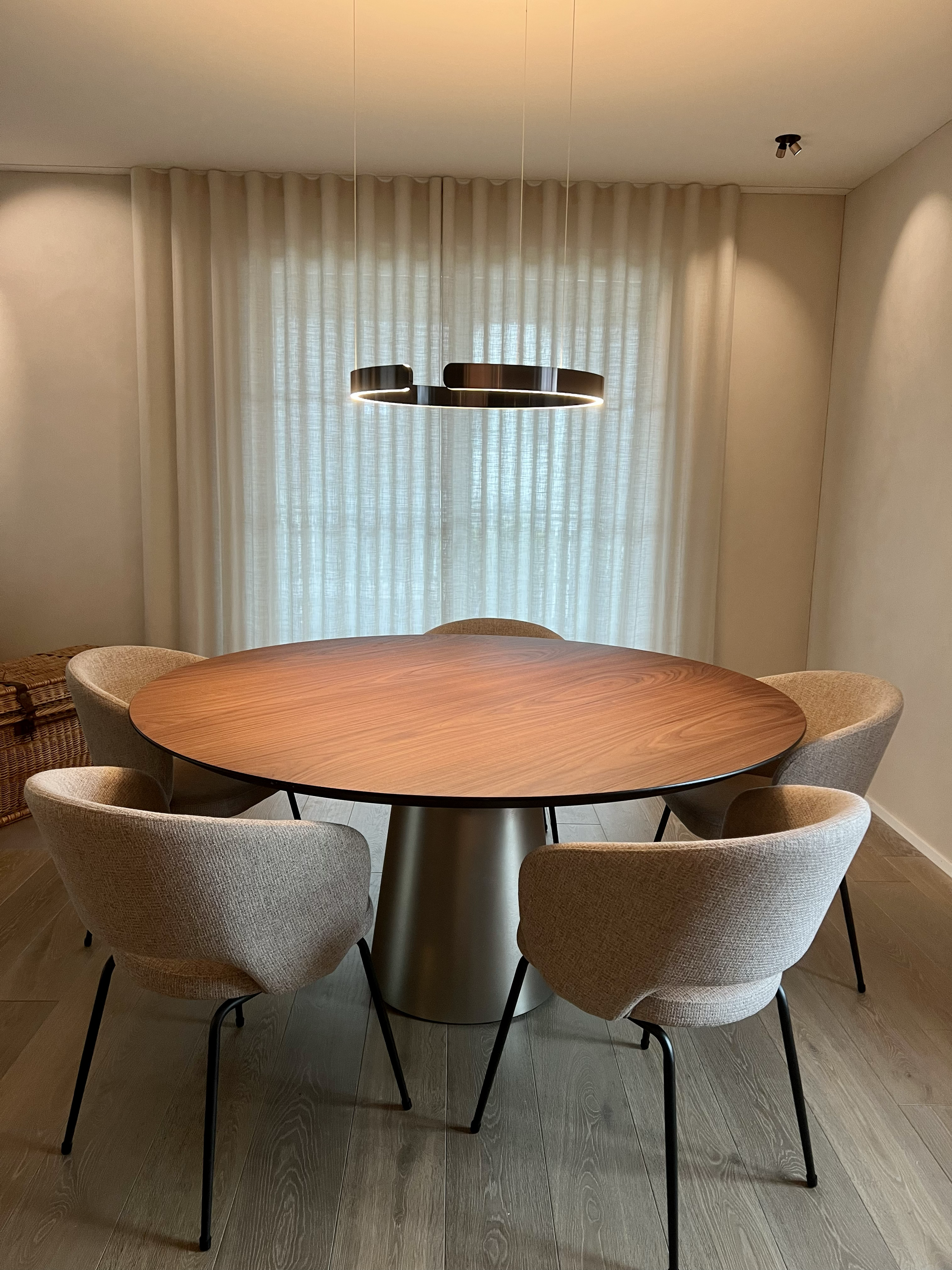 Ein runder Holztisch mit vier beige Stoffstühlen in einem hellen Esszimmer mit Vorhängen und moderner Hängelampe.