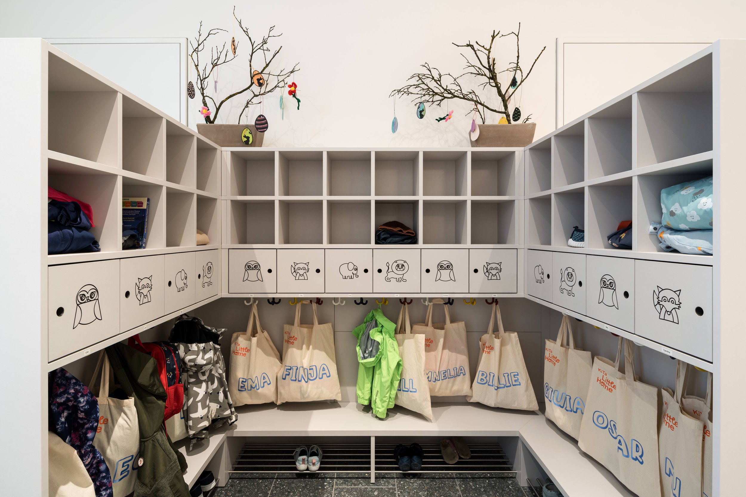 Kinderjacken, Schuhe und Taschen in einem organisierten Eingangsbereich mit weißen Schränken und dekorativen Zweigen auf den oberen Regalen.