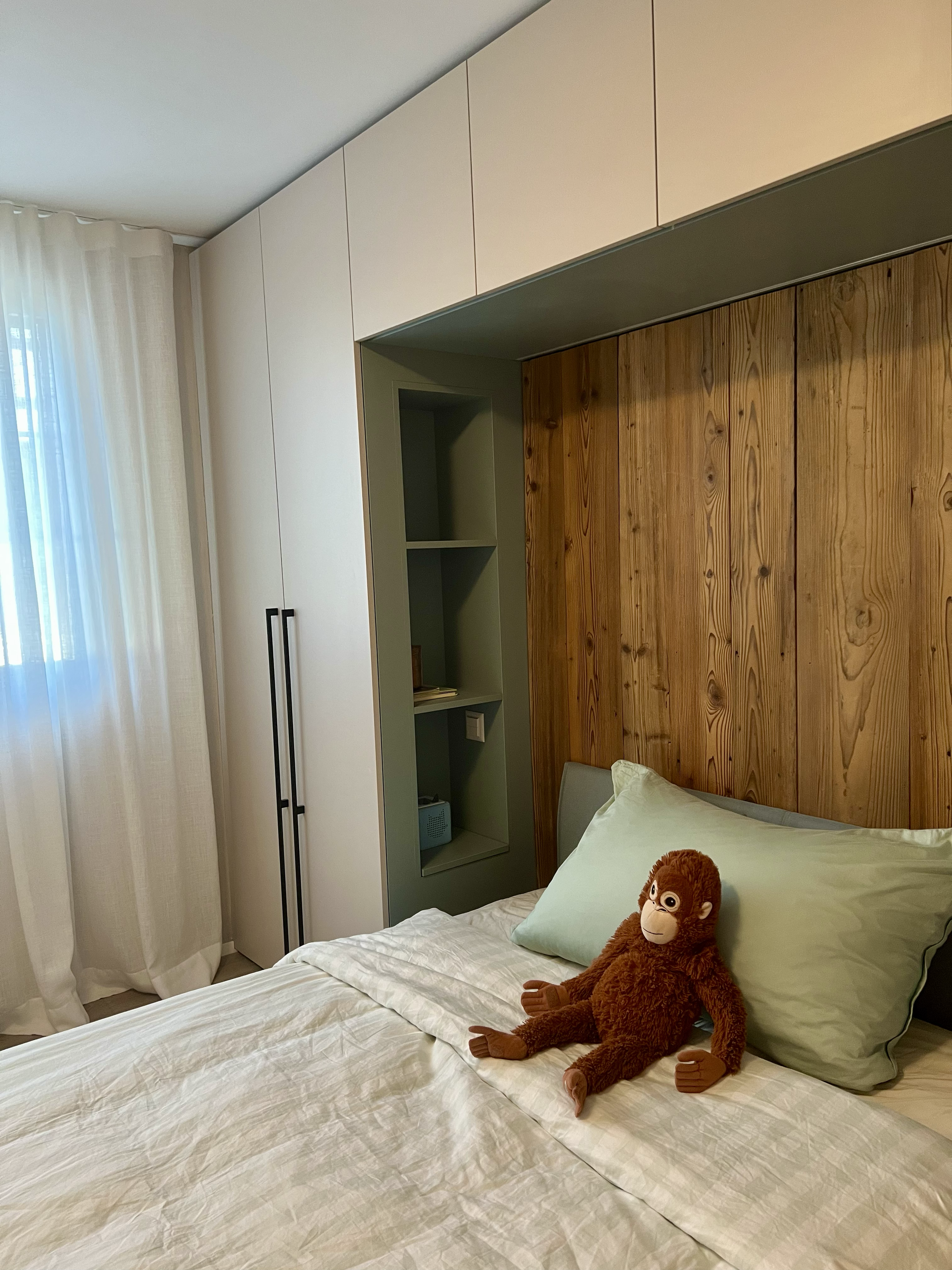 Auf einem Bett liegt ein Plüschtier eines Affen mit braunem Fell, weißen Augen und einem freundlichen Gesichtsausdruck, im Schlafzimmer mit Holz- und weißen Wandelementen.