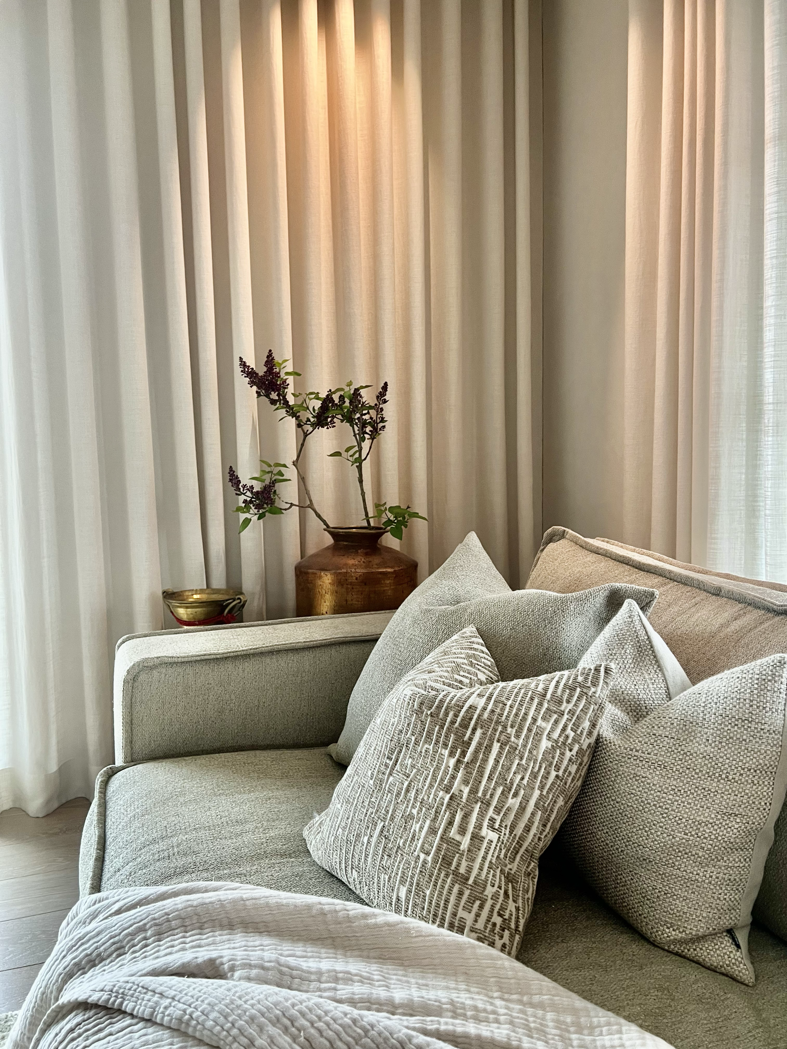 Ein Sofa mit mehreren Kissen in einem gemütlichen Wohnzimmer, im Hintergrund sind weiße Vorhänge und eine Vase mit Pflanzen auf einem Tisch