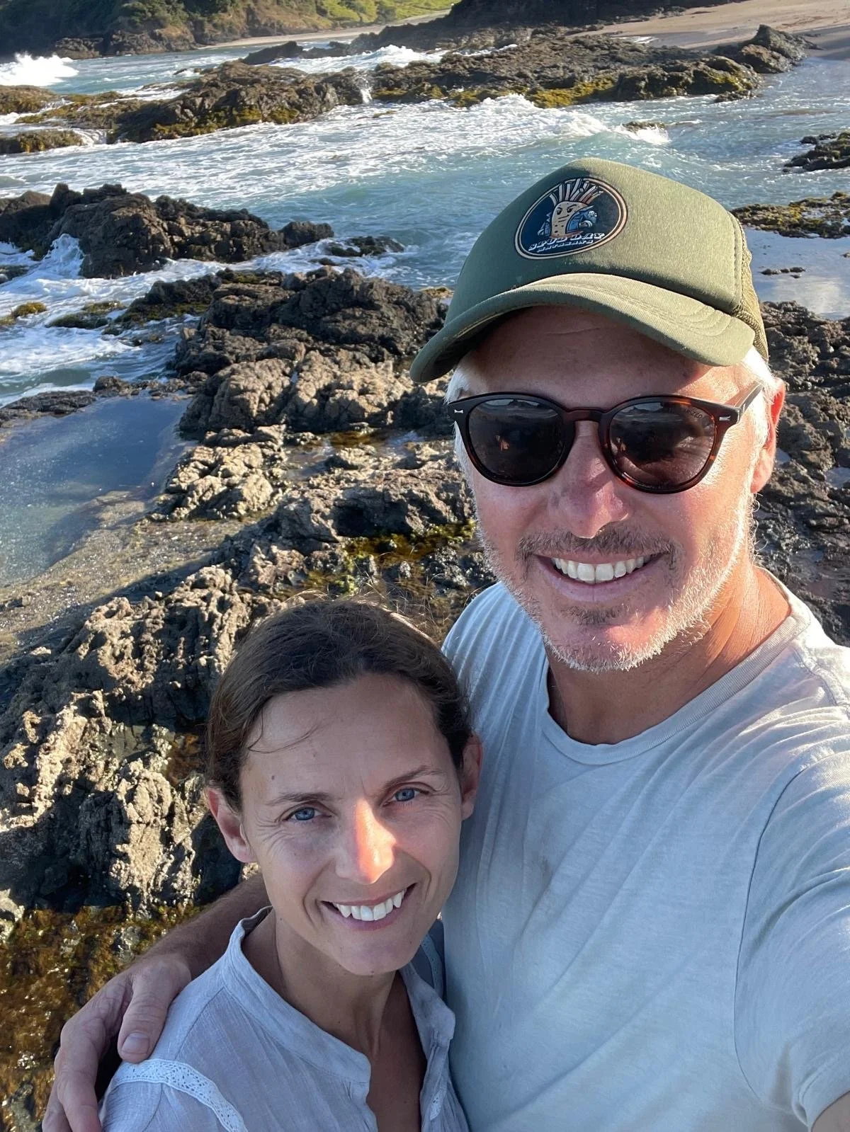 A smiling man and woman taking a selfie on a rocky shoreline with the ocean and waves in the background.