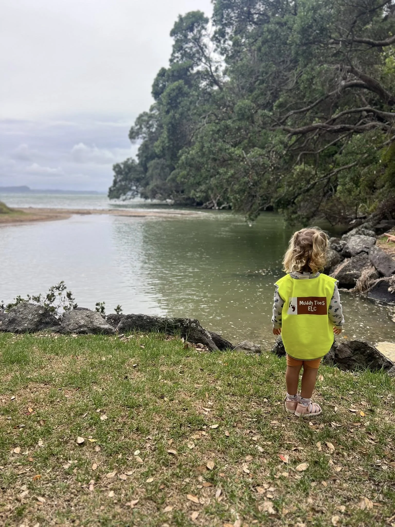 A young girl with curly blonde hair standing near a body of water, wearing a yellow vest that says "Muddy Toes ELC" on the back, looking out over the water in a natural outdoor setting with trees and rocks.