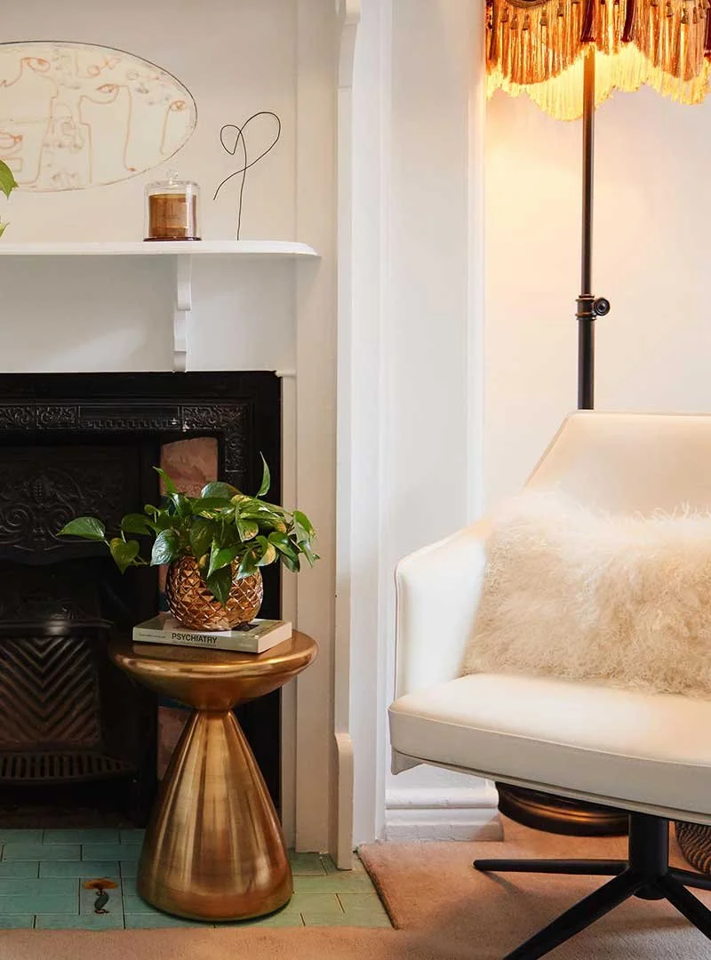 a cosy warm light up room with a white swivel chair and fluffy pillows that sit ontop of the chair. There is a brown coloured lamp and yellow and mustard coloured lamp shade. There is a fireplace with a shelf above the fireplace with a candle