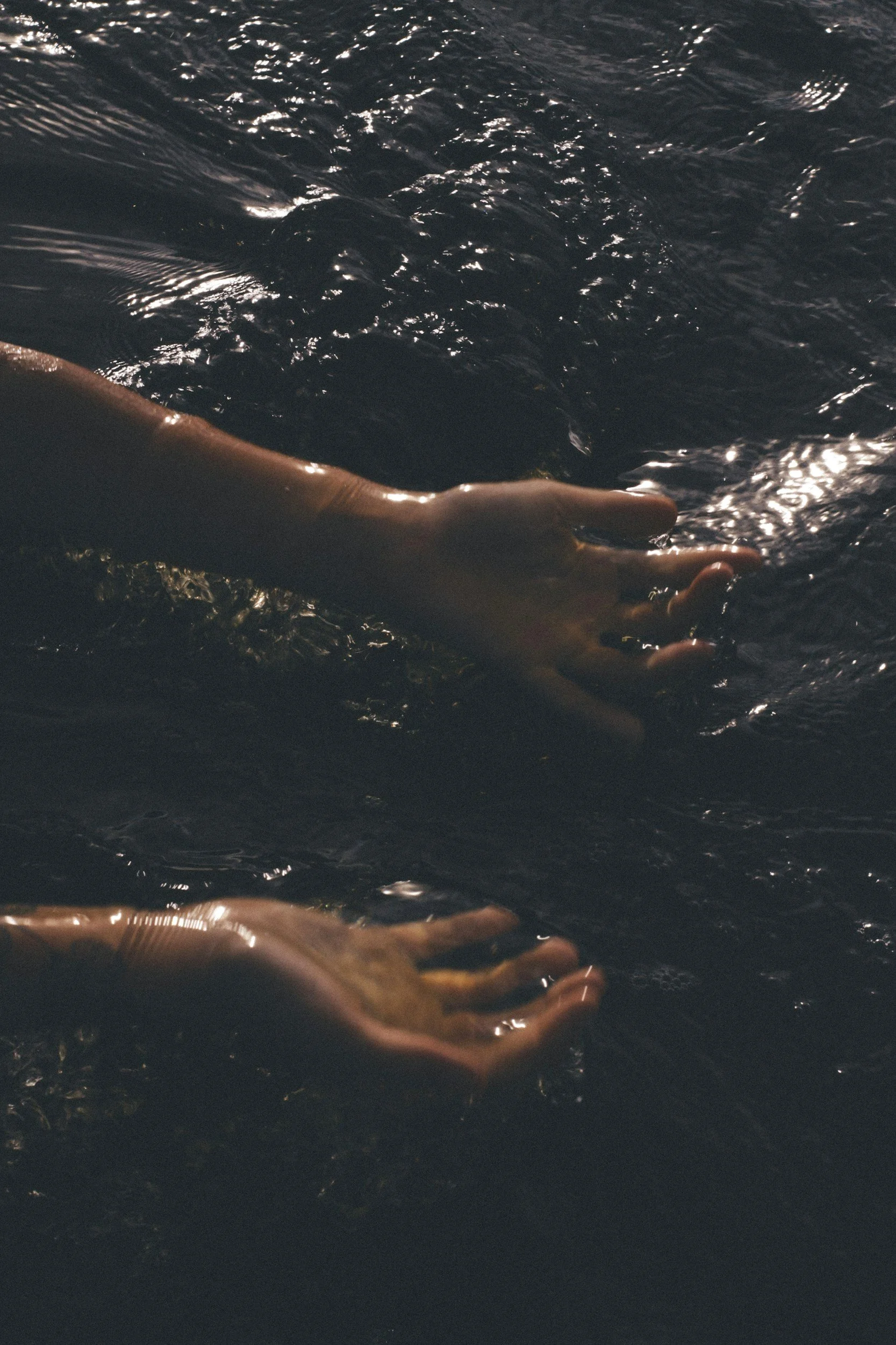 Two hands reaching into dark water, creating ripples.