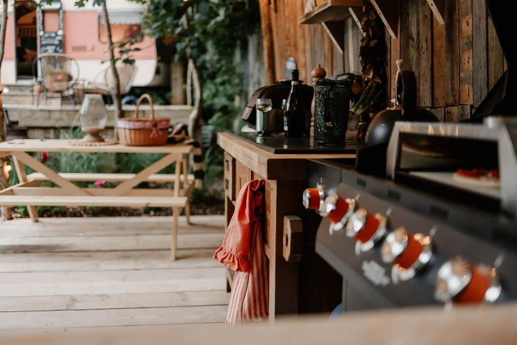 Outdoor kitchen with a black stove, wooden countertops, and various bottles and utensils, with a wooden table and benches in the background.