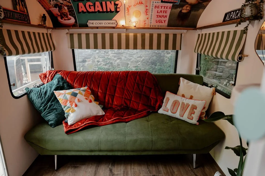Inside a cozy camper with a green sofa, colorful pillows, and a red blanket, set beneath large windows with striped valances showing a scenic outdoors.
