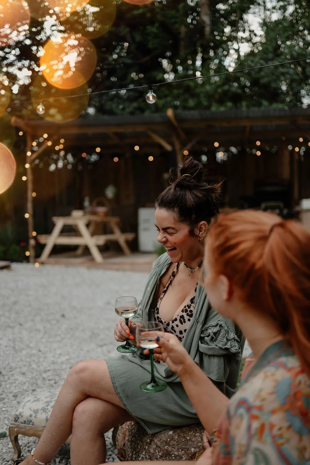 Two women sitting outdoors at a gathering, laughing and holding wine glasses, with string lights and a wooden structure in the background.
