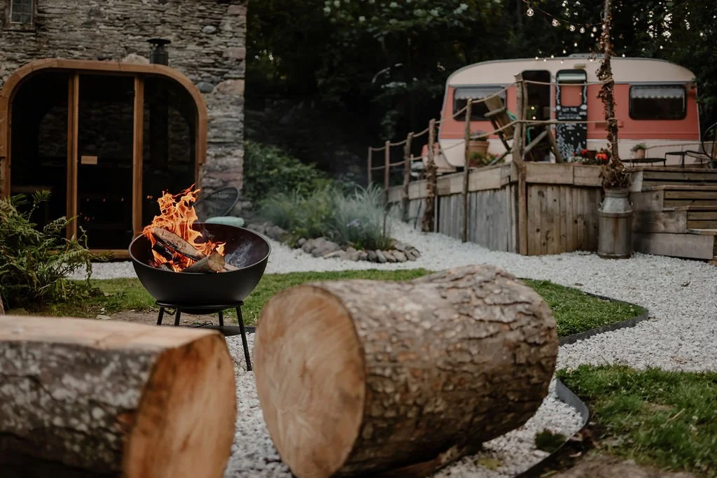 Backyard with a fire pit, logs, a small garden shed, and a vintage camper trailer with string lights and potted plants.