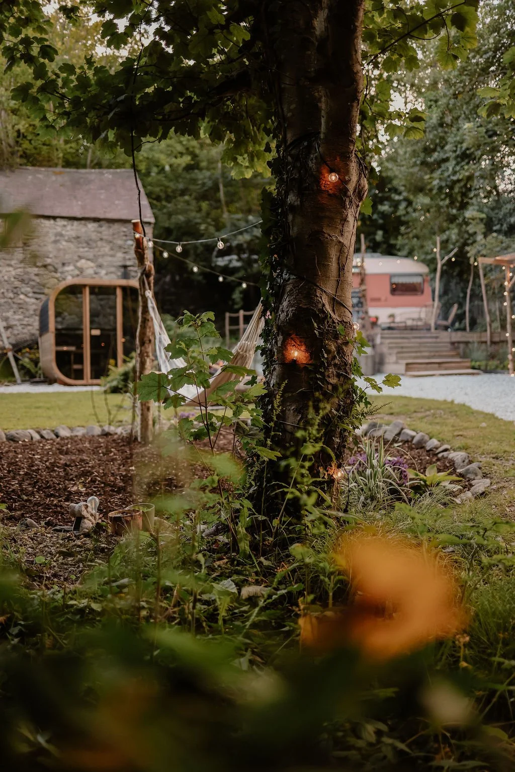A backyard garden with a tree wrapped in string lights, a stone border around the flower bed, and a hammock in the background. There is a small stone building and a vintage camper trailer.