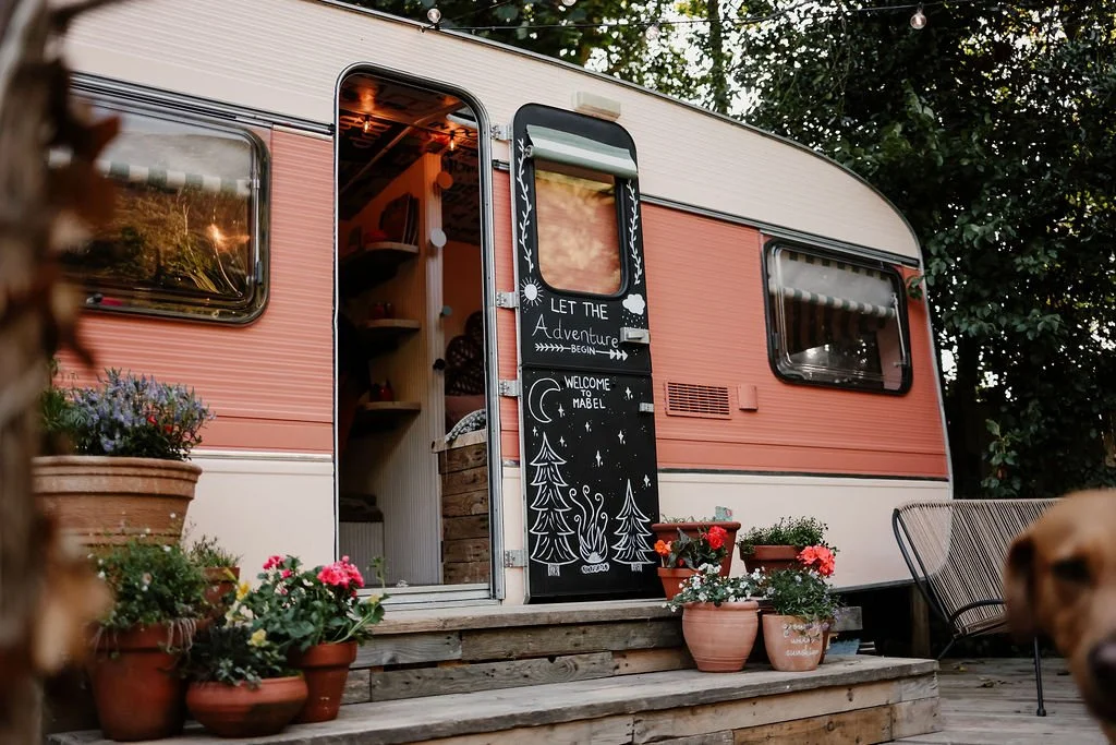 A vintage camper trailer with a pink and white exterior, decorated with potted flowers on a wooden porch. The camper's door has a chalkboard with whimsical drawings and texts that read 'Let the adventure begin' and 'Welcome to Mabel.' The interior vi