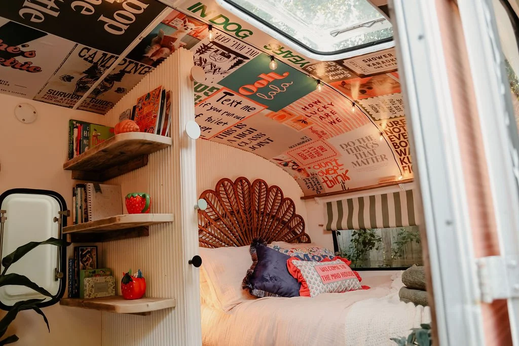 Cozy bedroom with a bed featuring decorative pillows and a rattan headboard, surrounded by decorated walls with books, small plants, and posters, and a window with striped blinds letting in natural light.