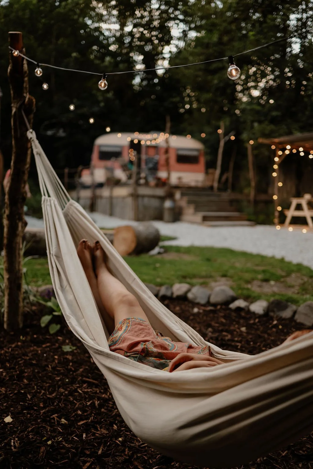 Person relaxing in a hammock outdoors at dusk, with string lights hanging overhead, a vintage trailer in the background, and a cozy outdoor setting with logs and string lights.