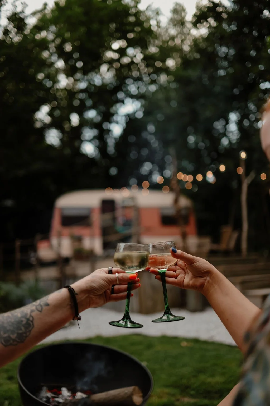 Two people toasting with glasses of wine or champagne outdoors during evening, with blurred background of trees, string lights, and a camper.
