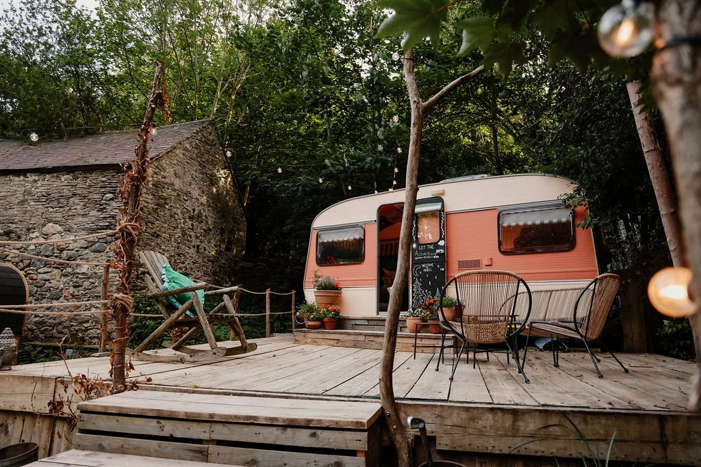 A cozy outdoor deck area with a vintage pink and white camper trailer, surrounded by lush trees and string lights, featuring patio furniture and decorative potted plants.
