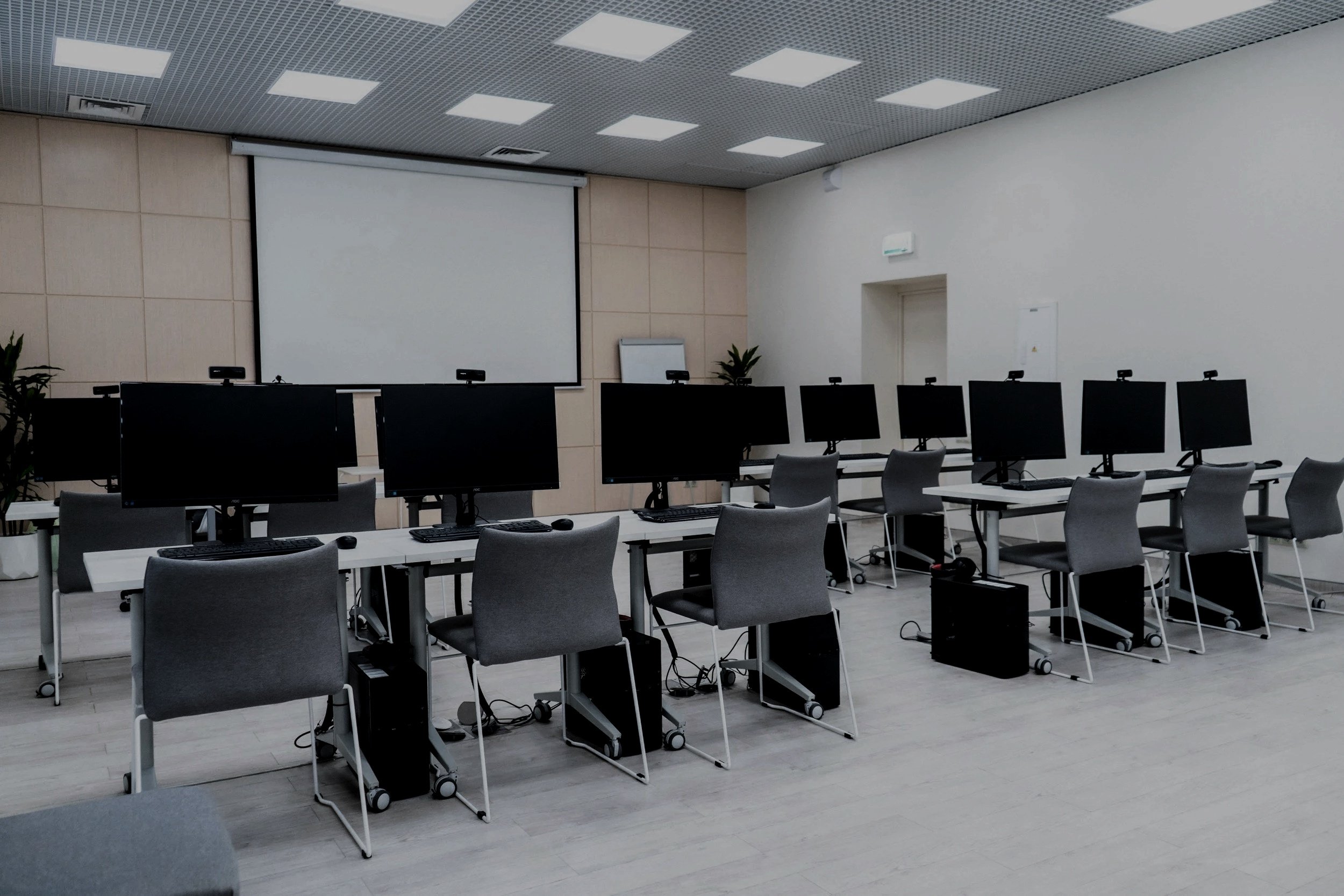 Empty computer training room with rows of desks, each with a computer monitor, keyboard, mouse, and gray chairs, white walls, and a large projection screen at the front.