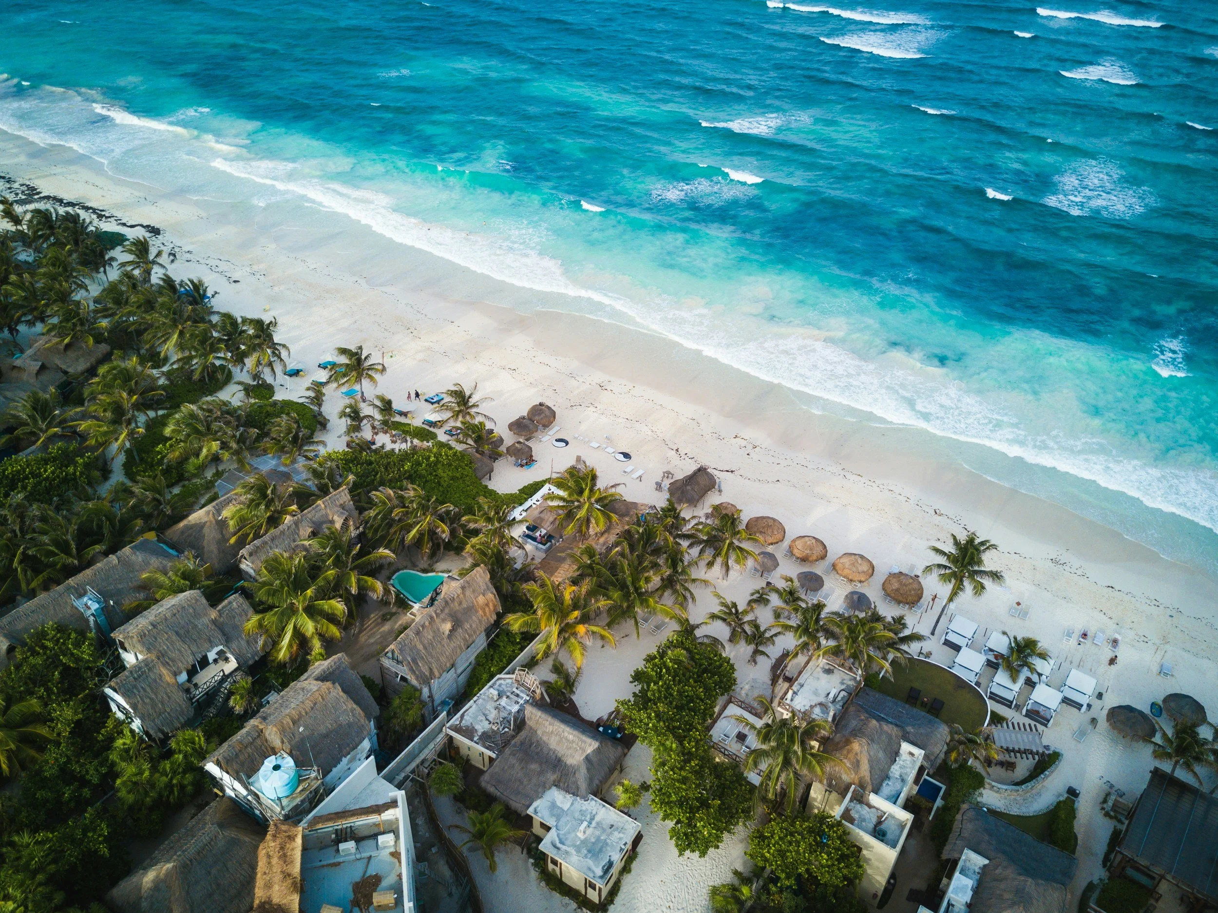 Aerial view of a tropical beach resort with thatched huts, palm trees, and lounge chairs along the white sandy shore, turquoise ocean waves crashing on the beach.