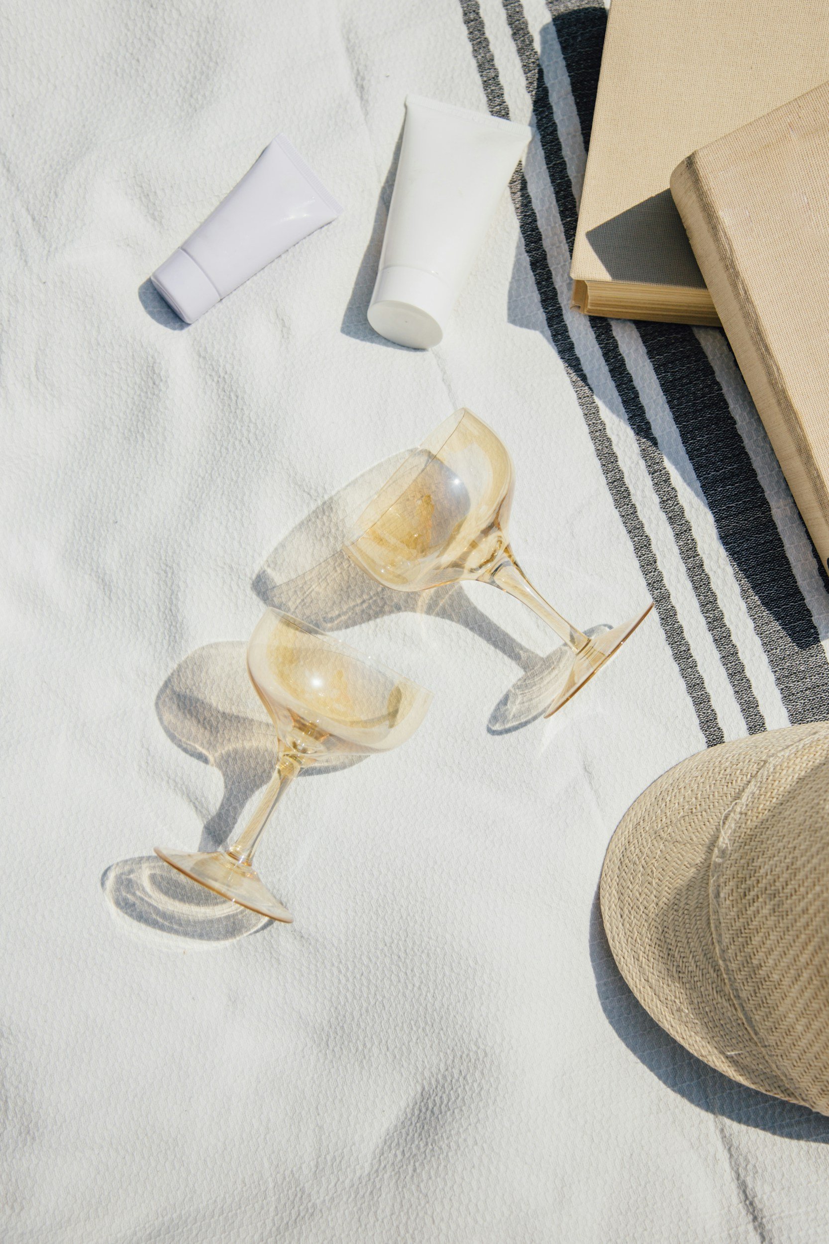 Two empty wine glasses, two white cosmetic tubes, two closed books, a woven hat, and a striped cloth on a white textured surface.