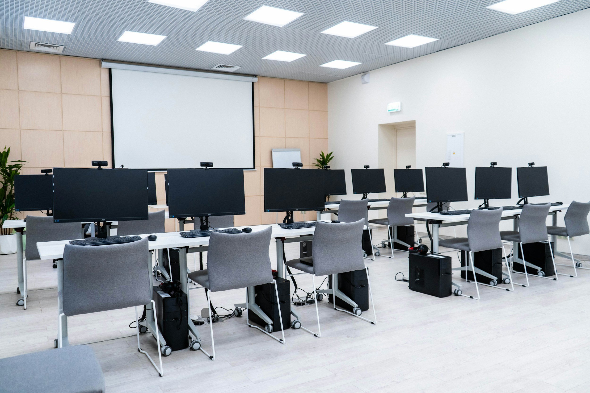 Empty computer training room with rows of desks, each with a computer monitor, keyboard, mouse, and gray chairs, white walls, and a large projection screen at the front.