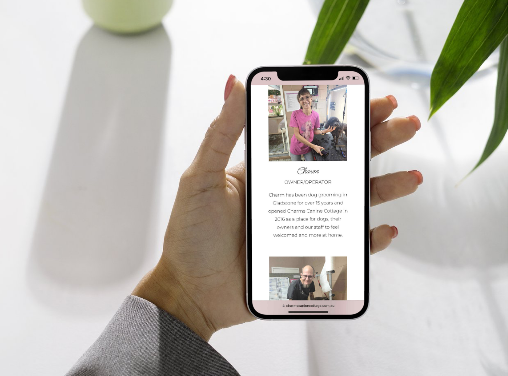 A person holding a smartphone displaying a profile for Charm, a dog grooming owner/operator at Charms Canine Cottage, with a photo of her smiling and grooming a dog at a grooming station.