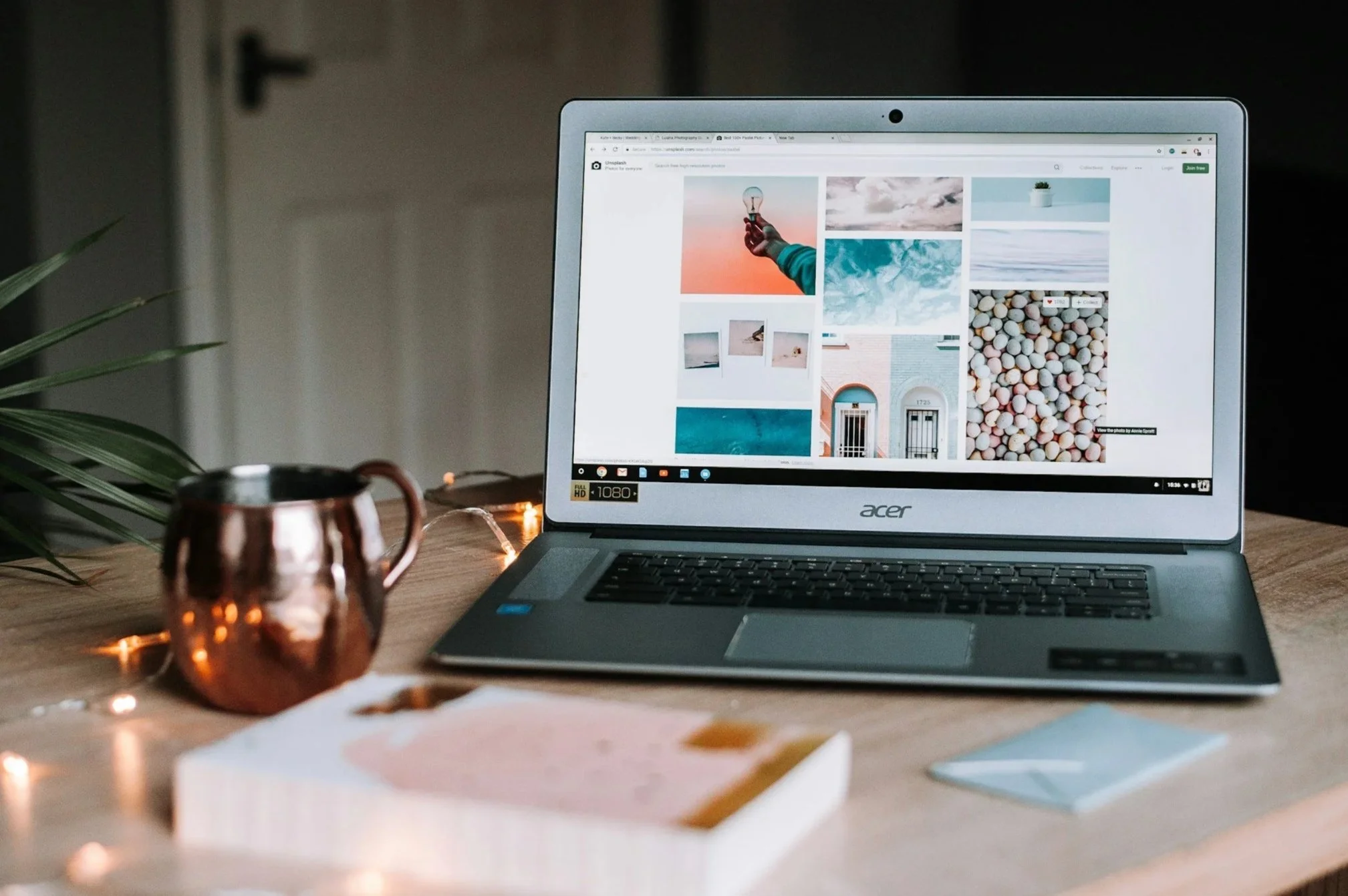 Open laptop on wooden desk displaying a collage of photos, with a copper mug, a small plant, a notebook, and a smartphone nearby, and fairy lights.