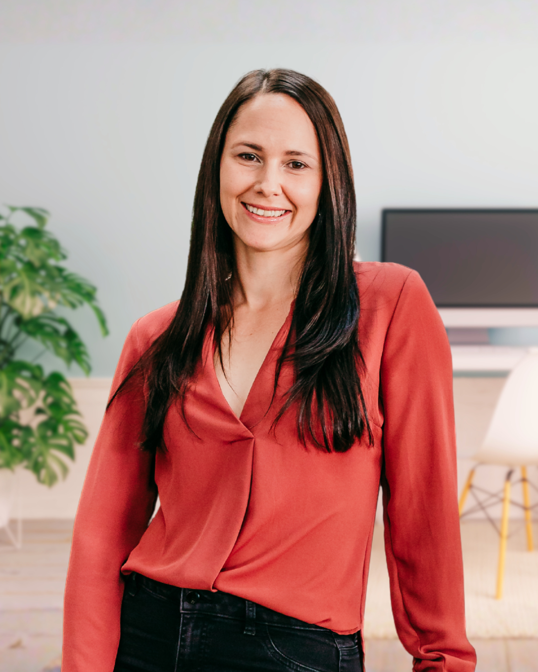A woman with long dark hair smiling in a bright, modern office with a green plant and a computer in the background.