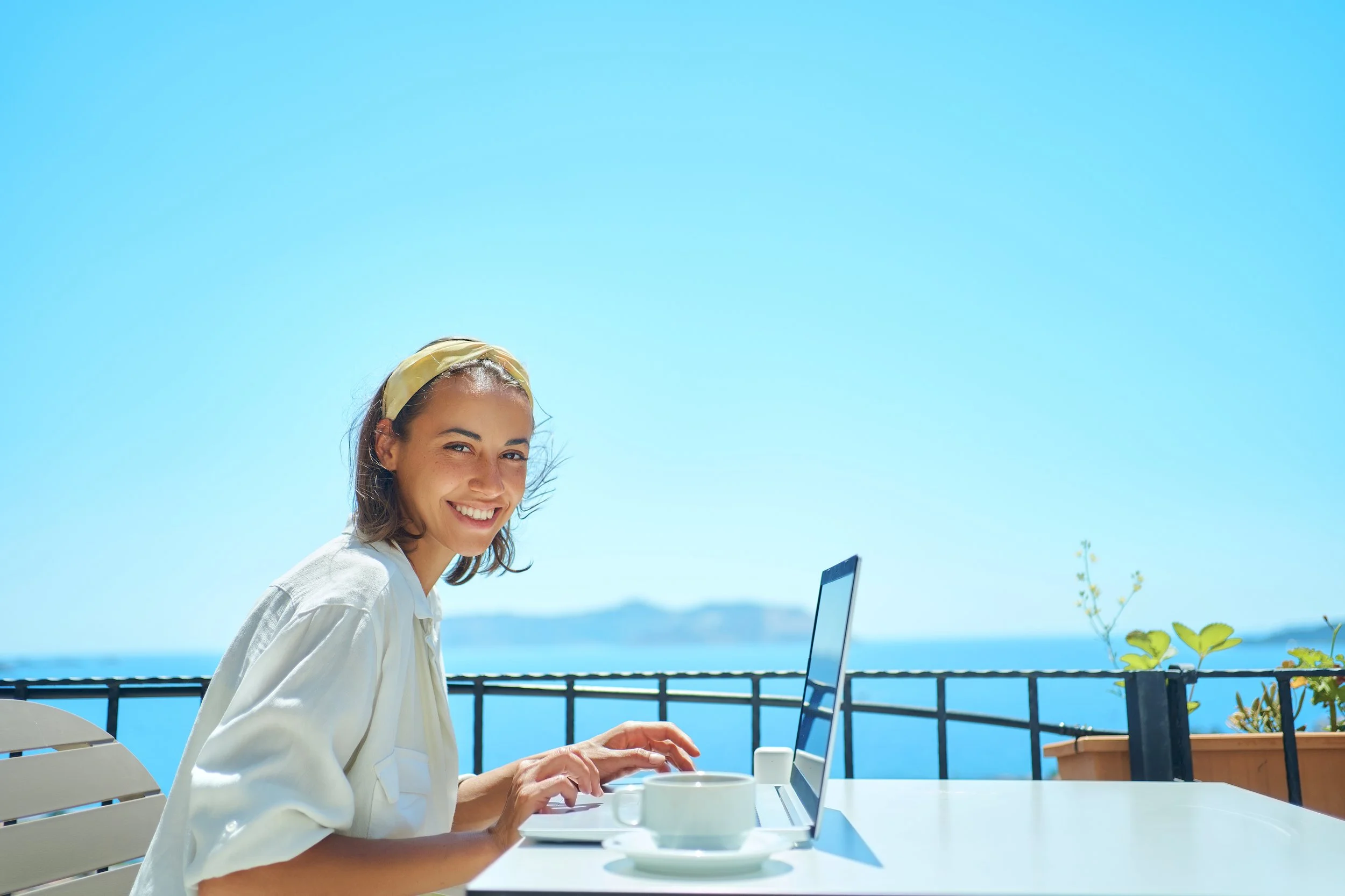 A woman sitting at an outdoor table with a laptop, with a view of the ocean and clear blue sky in the background.