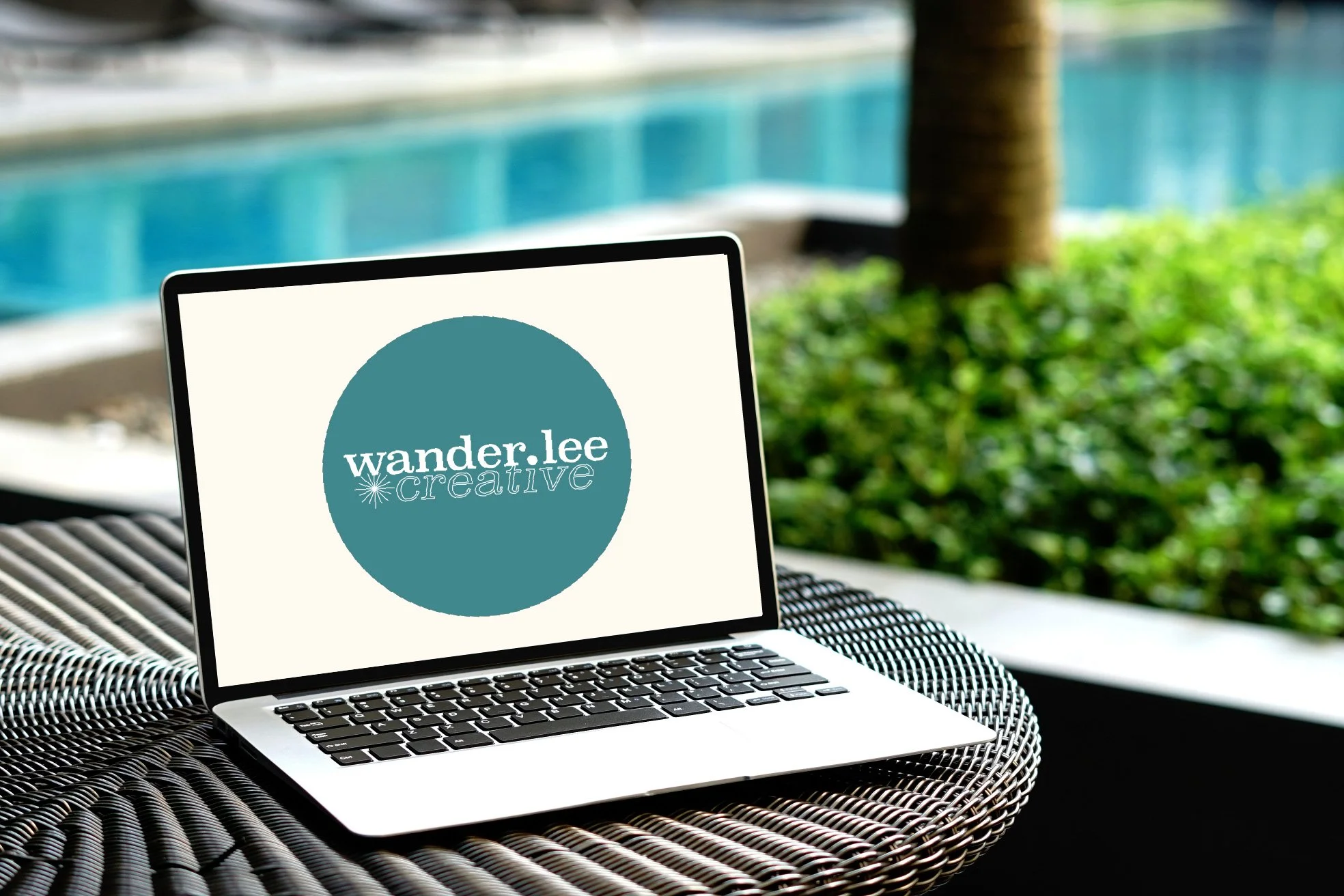 A silver laptop with a black keyboard displaying a logo that says "wander.lee creative" in white text on a teal circle, set on a woven outdoor table near a pool with green foliage in the background.