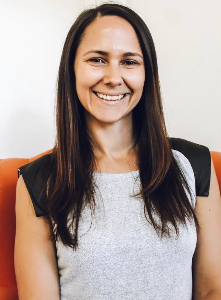 A woman with long brown hair smiling while sitting on an orange chair, wearing a sleeveless top with black shoulder accents.