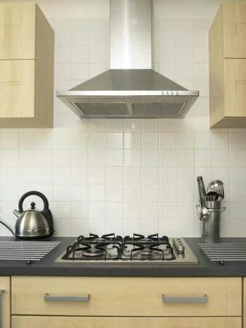 Kitchen stove with four burners, a stainless steel kettle, and a range hood, surrounded by beige cabinets and white tiled wall.