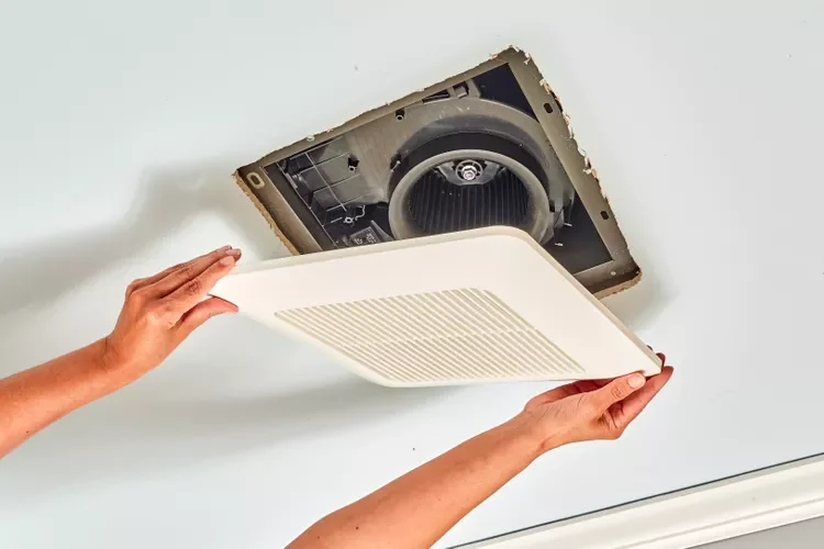 Person installing or replacing a ceiling vent cover, with the vent fan exposed in the ceiling opening.