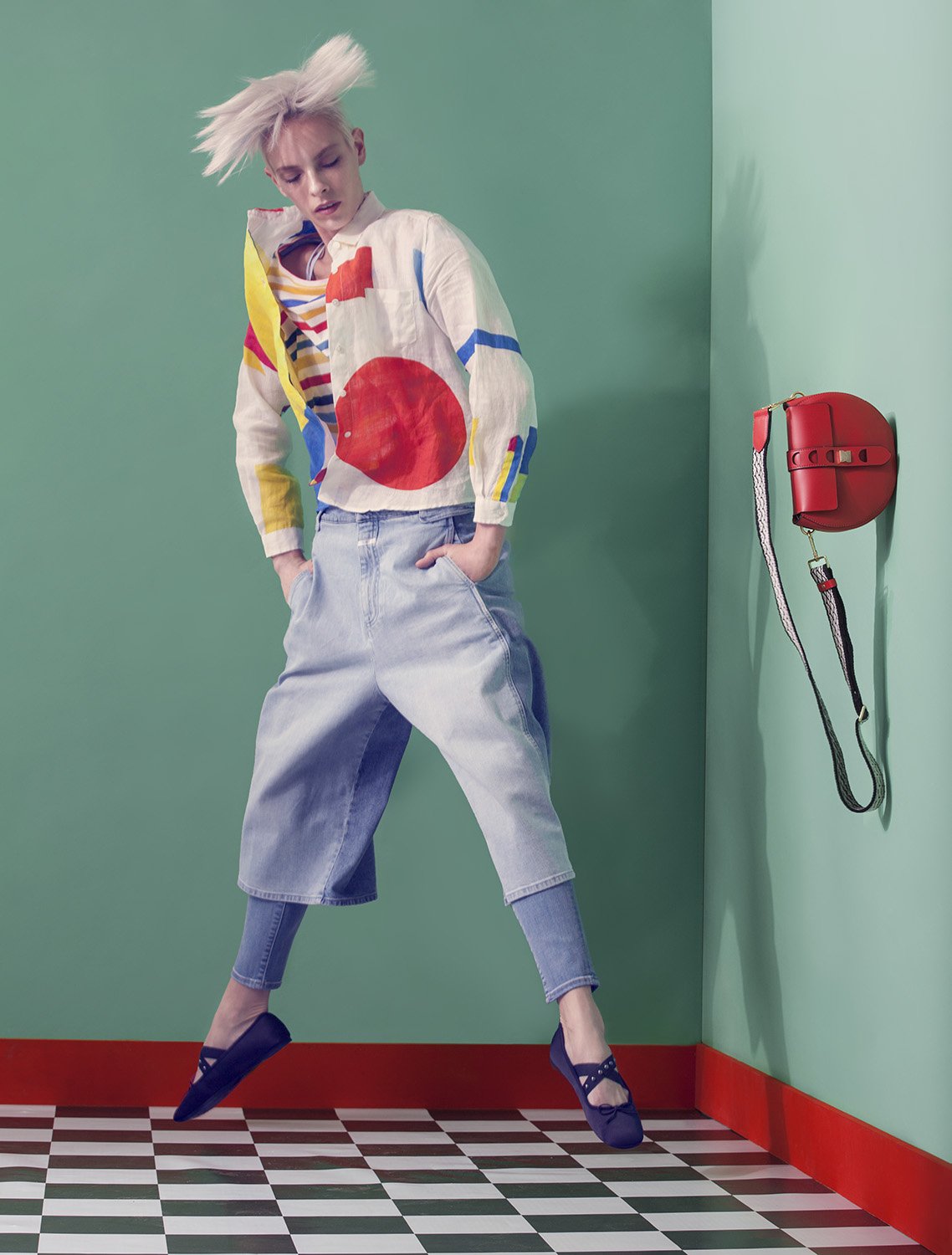 Editorial fashion image featuring model mid-jump against mint green corner wall with graphic shirt and hanging red handbag, set design and prop styling by Steffi Bühlmaier, photography by Christina Sandrock for Jolie, 2016
