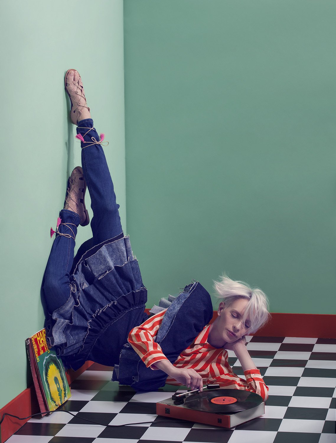 Editorial fashion image featuring model lounging upside down while playing vinyl record on checkerboard floor, set design and prop styling by Steffi Bühlmaier, photography by Christina Sandrock for Jolie, 2016