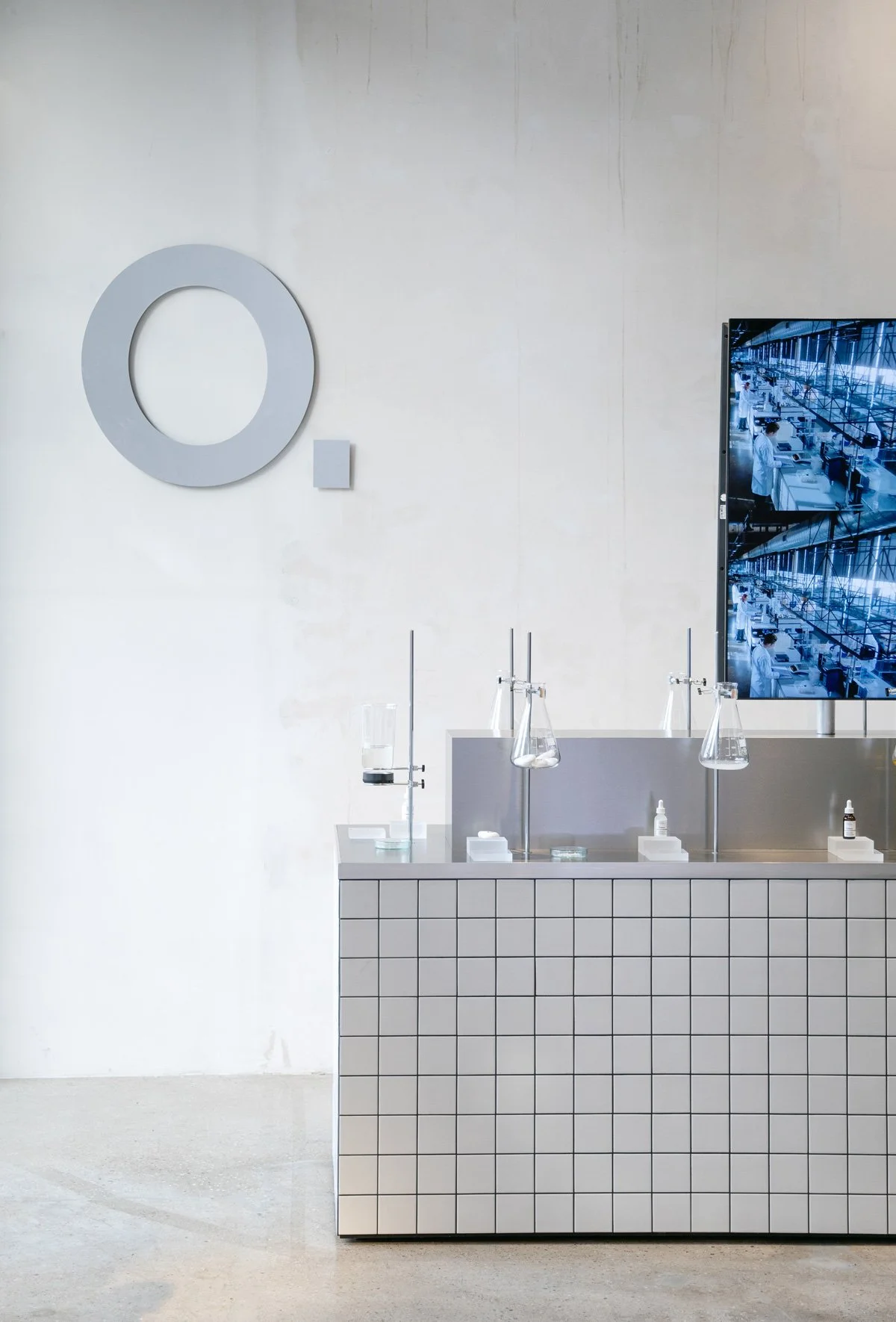 Minimal showroom installation with tiled counter, suspended flasks and digital screens referencing scientific aesthetics, display design and props styling by Steffi Bühlmaier for The Ordinary x Highsnobiety temporary showroom, event production by PS: