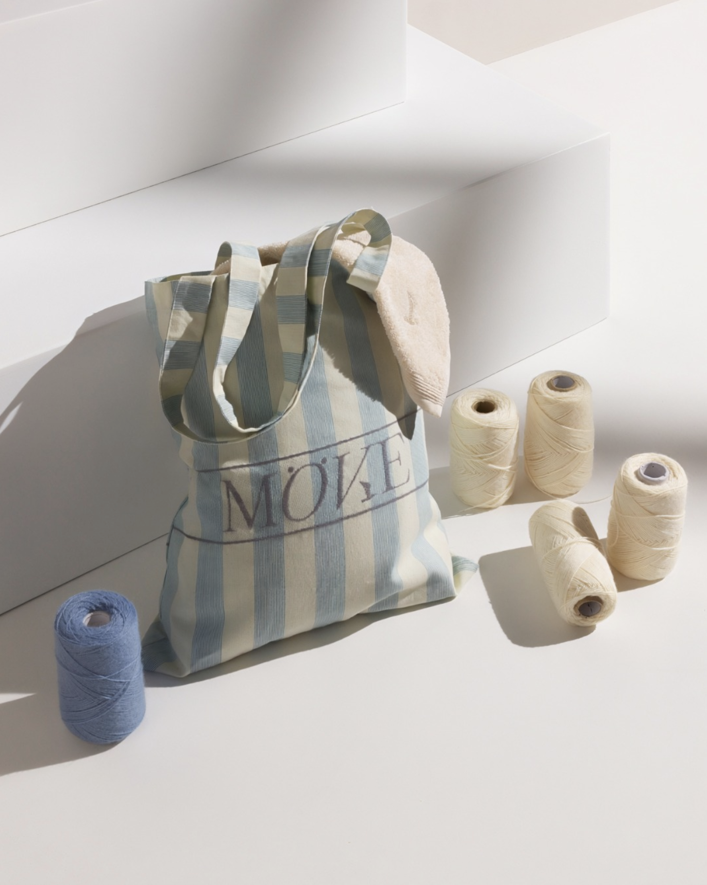 Still life featuring a bag with MÖVE logo stiching and and thread cones placed in front of white pedestrals, set build and props styling by Steffi Bühlmaier, photography by Michael Tewes, creative direction by Studio Kühn for Möve, 2025