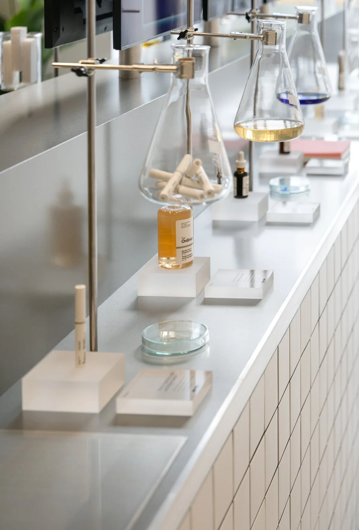 Close-up of laboratory-style glass vessels presenting skincare products on tiled display counter, display design and props styling by Steffi Bühlmaier for The Ordinary x Highsnobiety temporary showroom, event production by PS:KAAS, photography by J.K