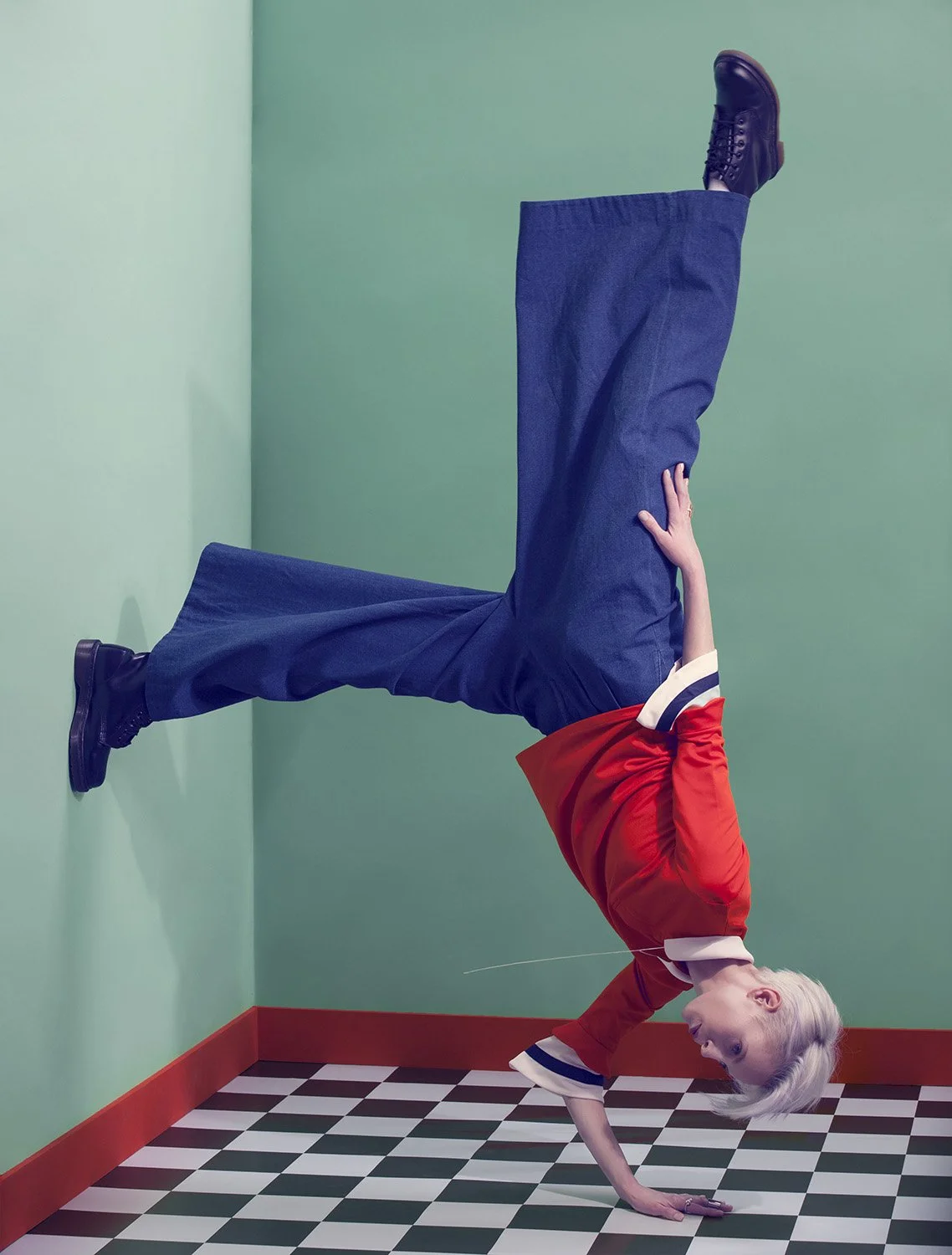 Editorial fashion image featuring model performing inverted handstand pose against wall in red top and wide trousers, set design and prop styling by Steffi Bühlmaier, photography by Christina Sandrock for Jolie, 2016