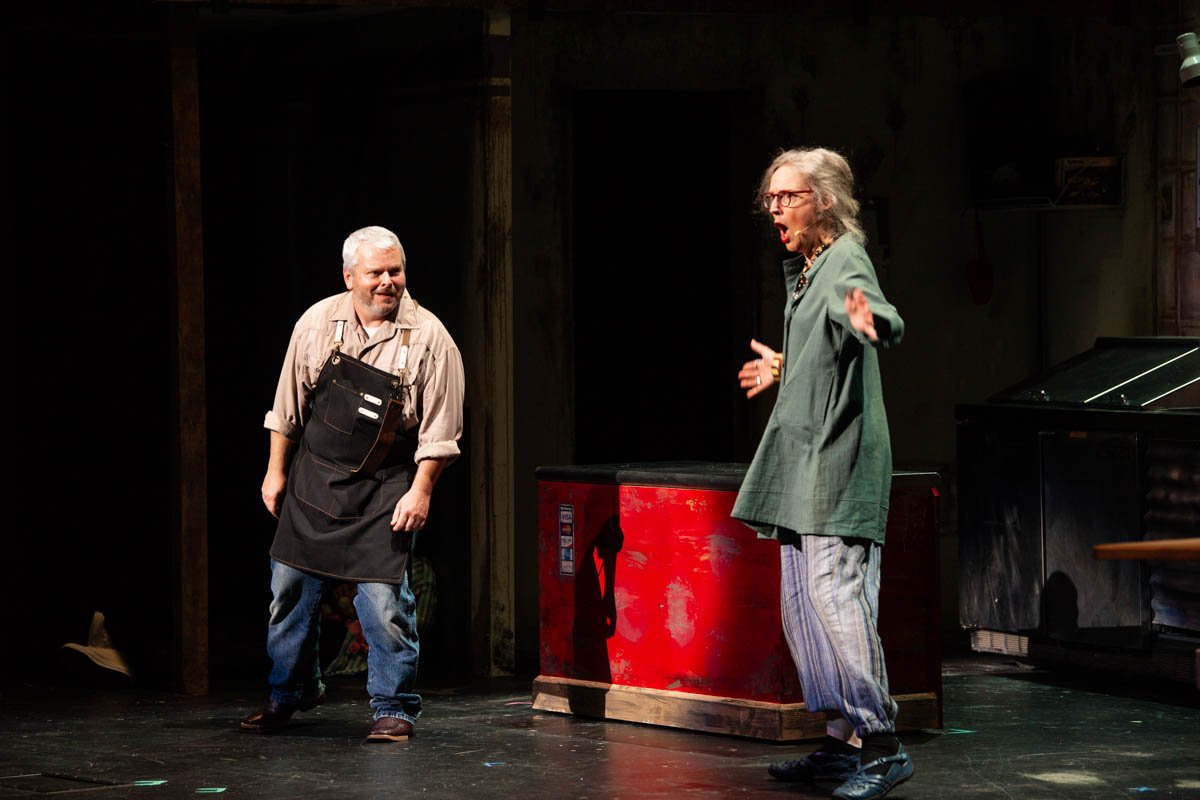 Two actors performing on stage, one man wearing an apron and jeans, and a woman with glasses wearing a green jacket and striped pants, engaged in a theatrical scene.
