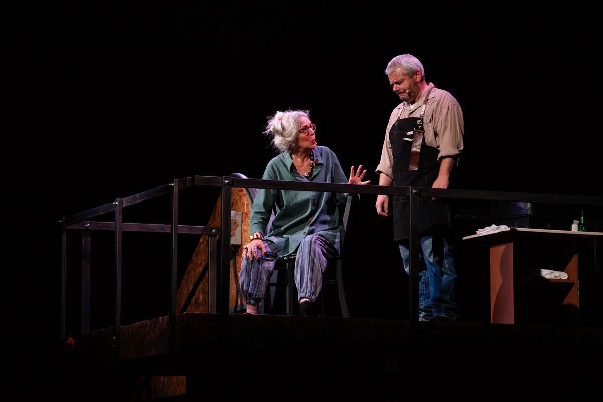 An elderly woman with gray hair and glasses sitting on a chair and talking to a man standing next to her on a stage. The woman is wearing a green jacket and striped pants, while the man has gray hair and is wearing a beige shirt with black overalls. 