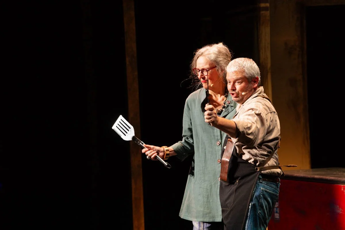 Two older actors on stage, one woman and one man, performing in a play. The woman is holding a spatula, and the man is pointing forward with a humorous expression.