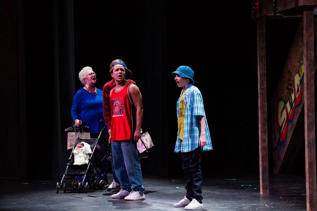 Three actors on stage performing in a play. The woman on the left has white hair, glasses, and is wearing a blue outfit; she is pushing a stroller. The young man in the middle is wearing a red sports jersey, a headband, and has a surprised expression