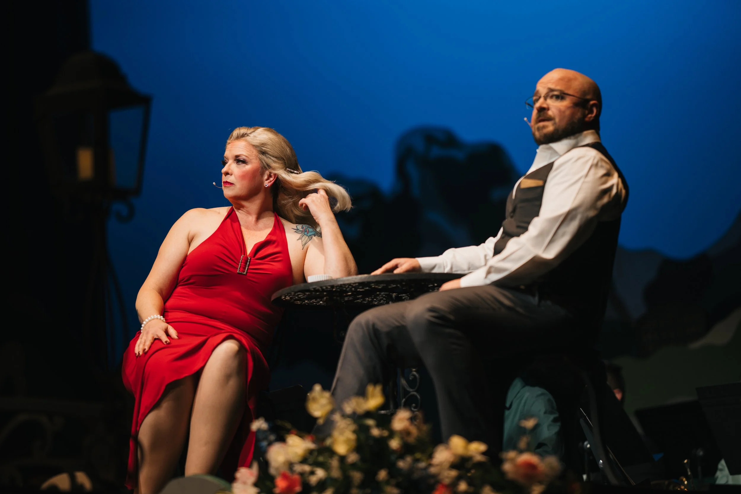 A woman in a red dress sitting at a table with a man in a white shirt and black vest, both wearing microphones, on a stage with a dark blue background and decorative flowers in the foreground.