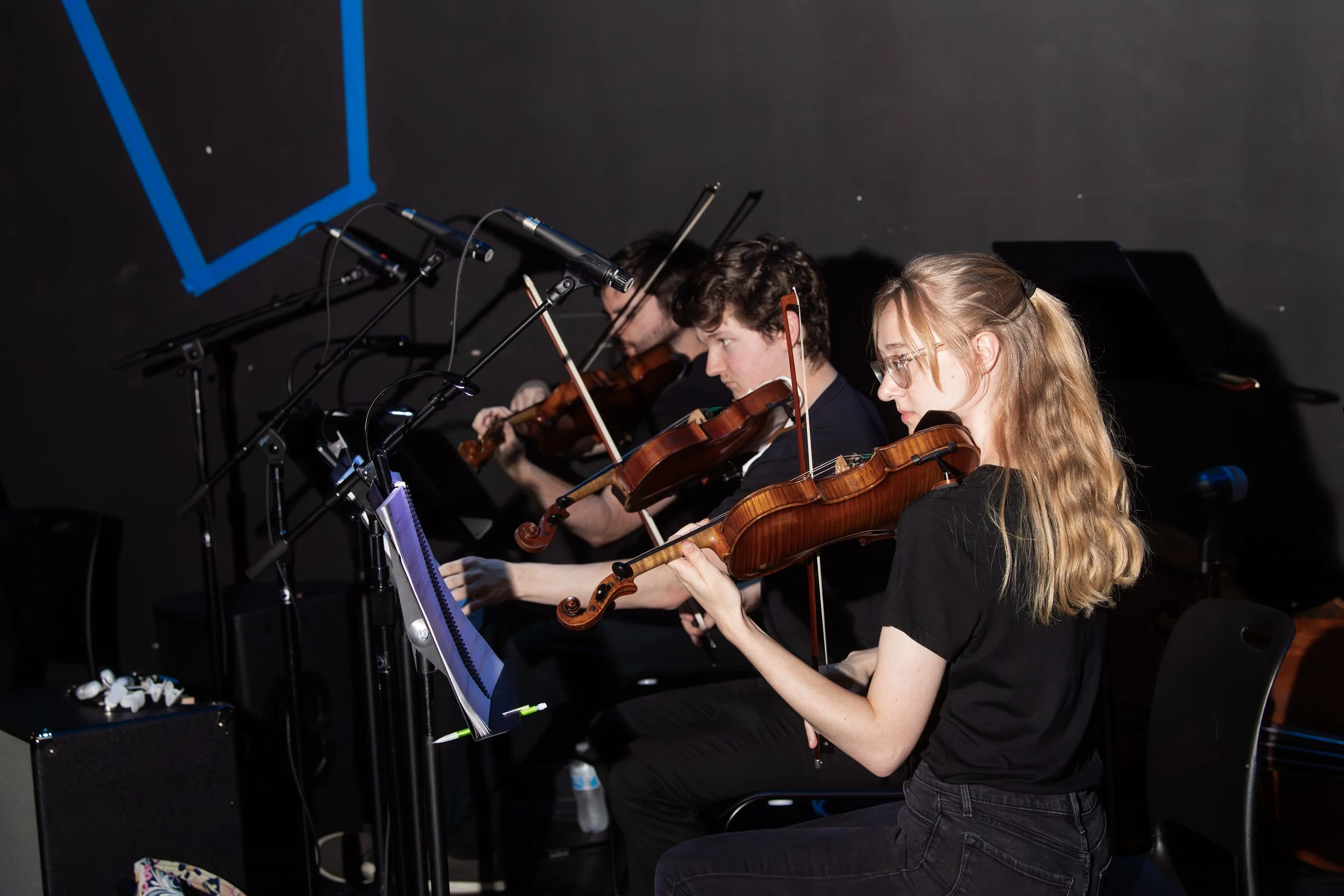 Four musicians playing violins on stage with music sheets, microphones, and a black background