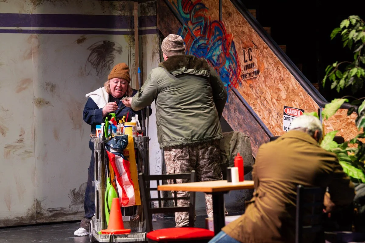 A woman with a brown beanie serving a man in military-style clothing at a colorful hot dog cart inside a sketchy-looking building.
