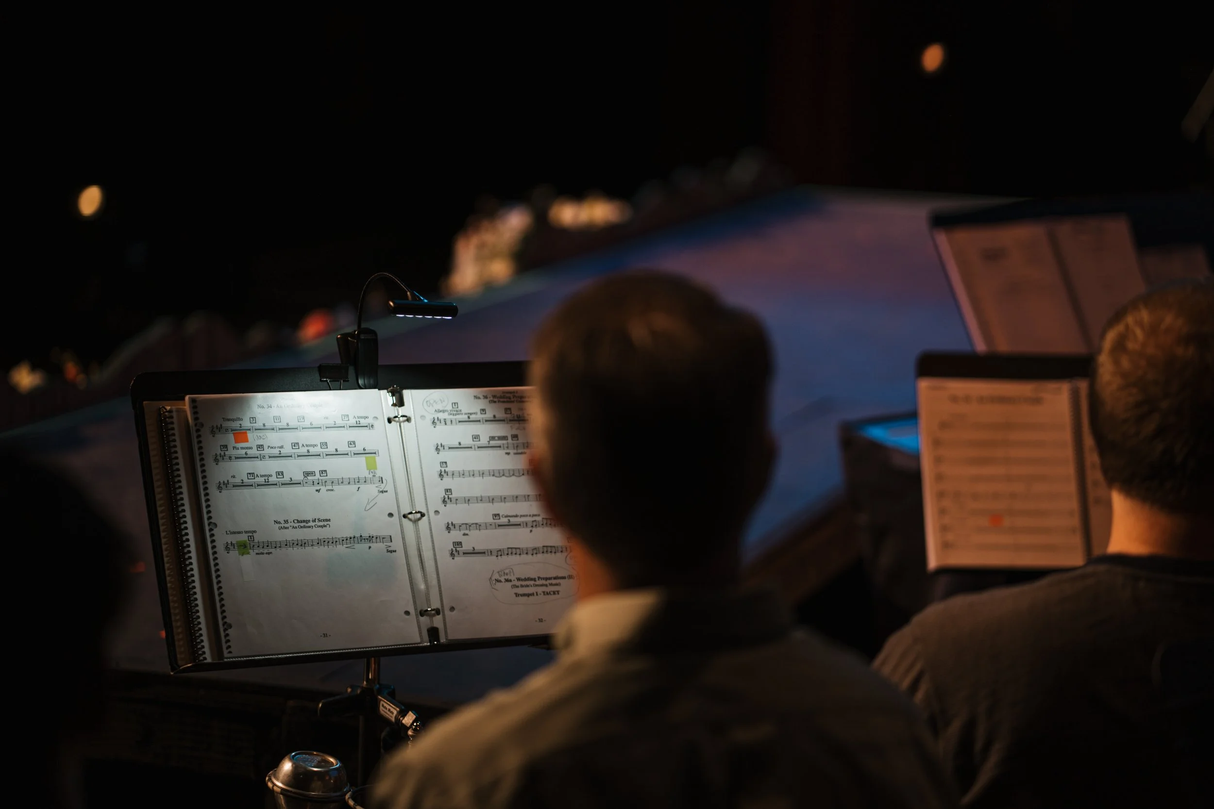 Musicians reading sheet music at night, illuminated by music stand lights, with a dark background.