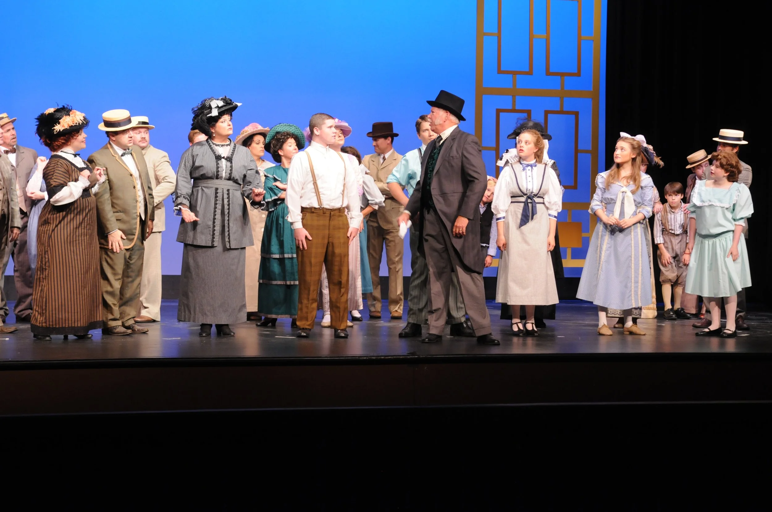 A theater stage with a cast of actors dressed in vintage 19th or early 20th-century clothing, engaged in a scene. The background is a plain blue backdrop with decorative wooden panels.