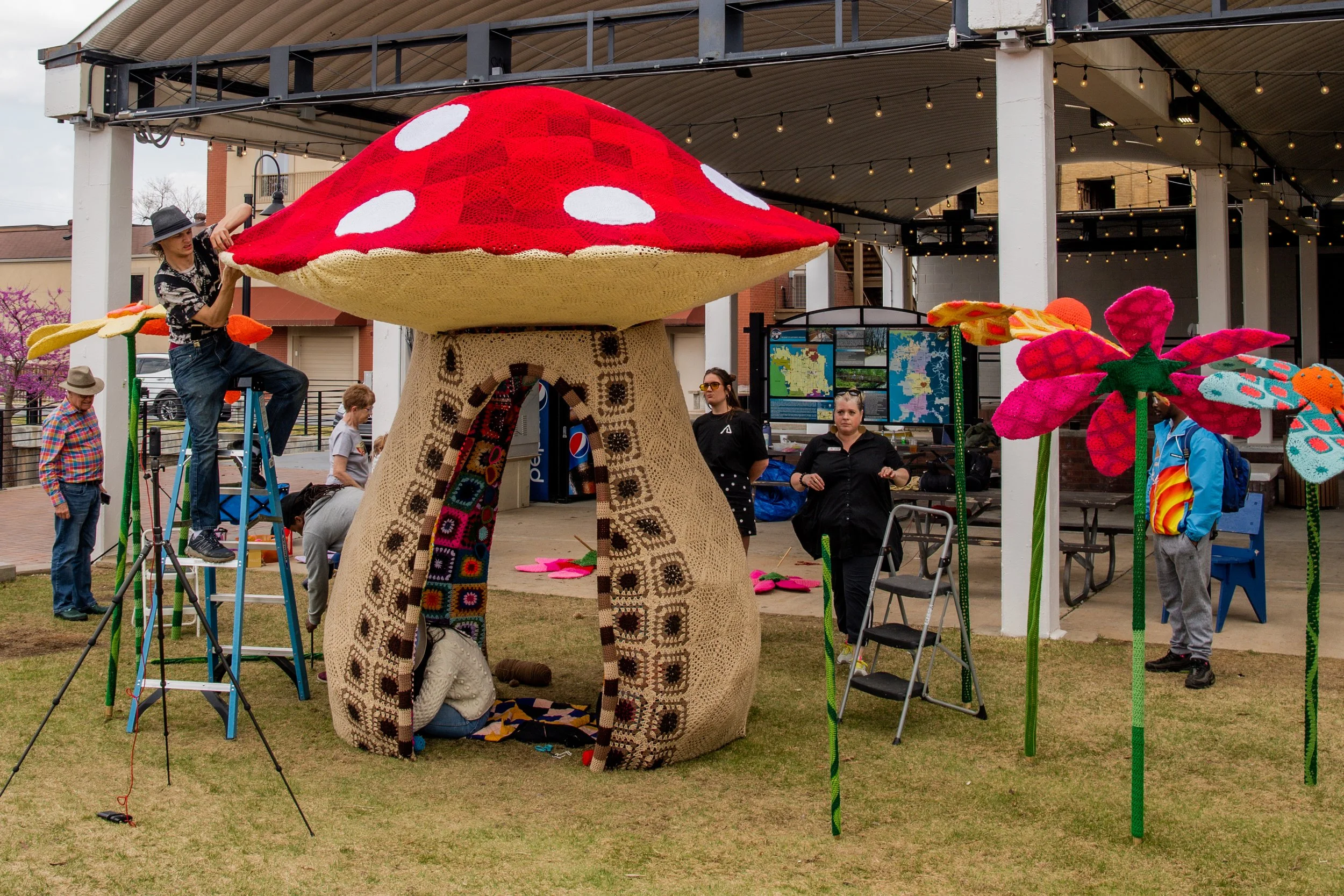People setting up large colorful crocheted mushroom and flowers sculptures on a grassy area near an outdoor shelter with string lights, with some individuals working on the sculptures and others observing.