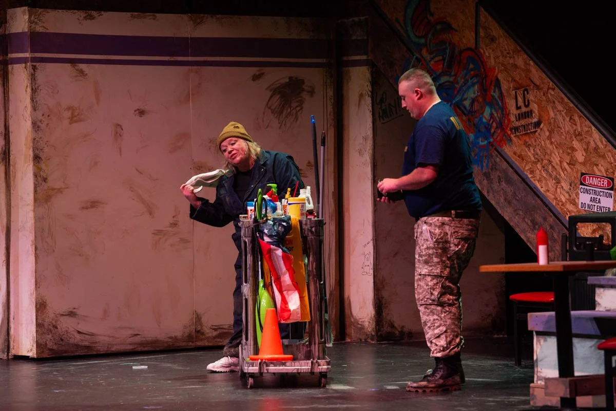 A woman in casual clothing holding a cloth, standing next to a cart filled with cleaning supplies and an orange traffic cone, with a man in camouflage pants looking down, inside a construction-themed setting.