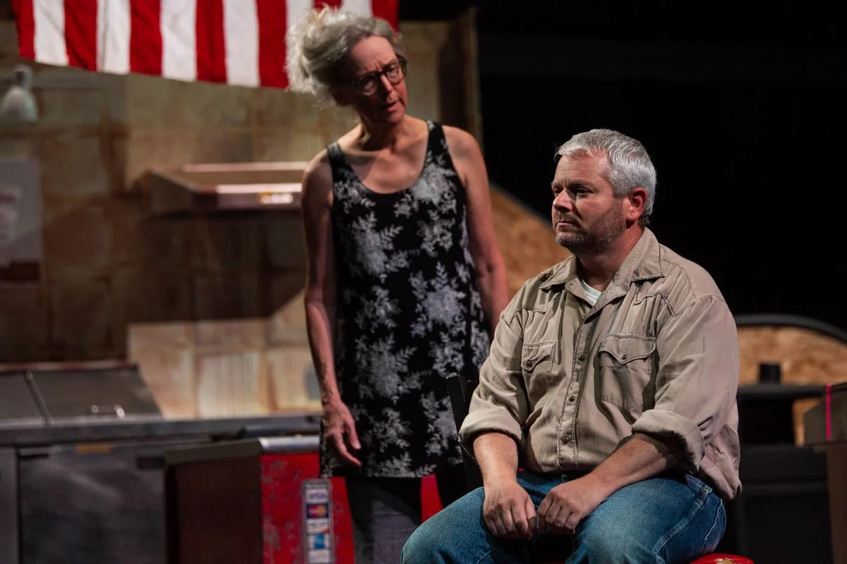 A woman with gray hair and glasses wearing a black and white floral dress stands behind a seated man with gray hair and a beard, wearing a beige shirt and jeans, in a room with a wooden background and an American flag hanging in the background.